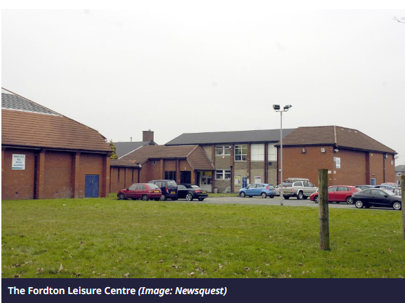 The Bands Name Sake A leisure centre building with cars parked outside in a cloudy setting.